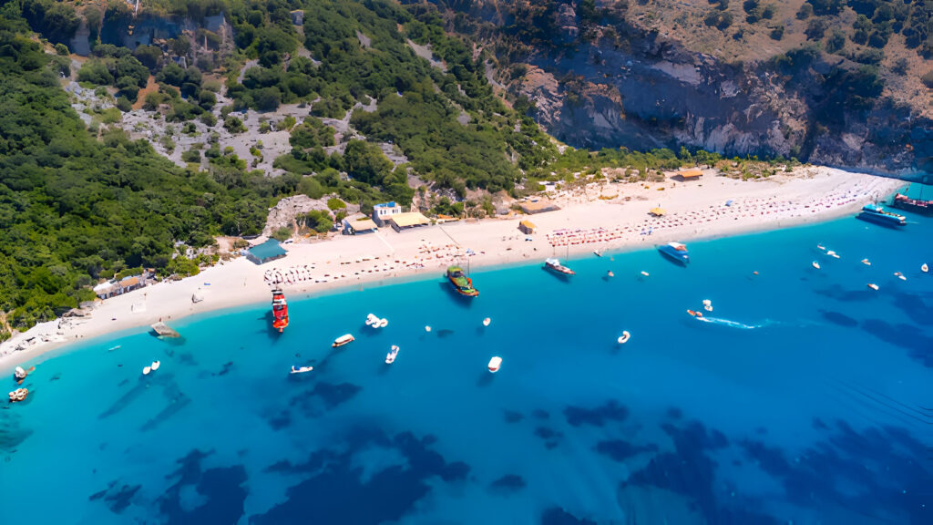 Renting a Car for the Albanian Riviera: Coastal Drives & Beach Stops 2 Aerial view of a stunning beach on the Albanian Riviera, featuring clear turquoise waters, boats anchored near the shore, and a sandy beach surrounded by lush green cliffs, perfect for renting a car for the Albanian Riviera to explore.