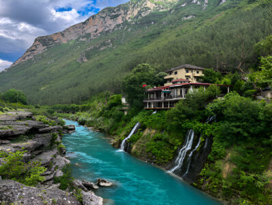 Picturesque waterfall and turquoise river near a charming mountain lodge in Valbona, one of Albania's hidden gems, ideal for a scenic road trip.