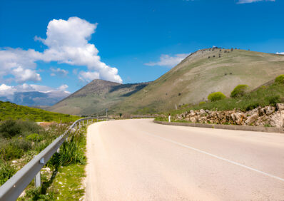 A picturesque road curving through the Albanian Riviera, flanked by green hills and a clear blue sky with fluffy white clouds, showcasing the natural beauty of the region.
