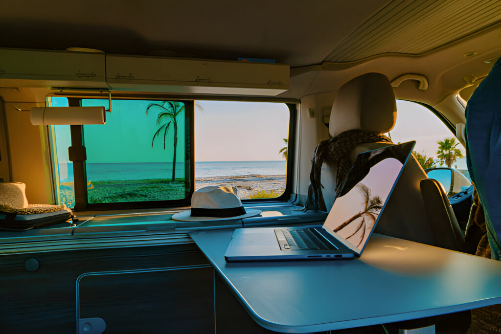 Interior of a van during an Albania road trip with a laptop on the table, a hat resting nearby, and a scenic beach view with palm trees through the window at sunset.